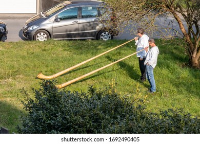 Flums, Switzerland - 03.04.2020: A Senior Is Given A Birthday Visit In The Form Of An Alphorn Duet While She Listens From The Balcony Of An Apartment Building In Flums.