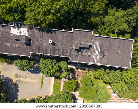 Flushing queens ny building complex featuring a flat dark roof with vents and skylights. Surrounded by lush green trees. An empty asphalt parking lot. And vibrant landscaping