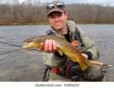 A fly fisherman posing with a Brown Trout with his fly rod and reel, before releasing the fish into the river