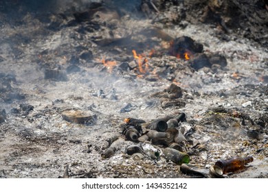 Fly Tipping And Arson. Shallow Depth Of Field Shows Flames, Smoke And Ash Blurred In The Distance Behind A Pile Of Beer Bottles And A Food Can Dumped As Trash In An Illegal Tip.