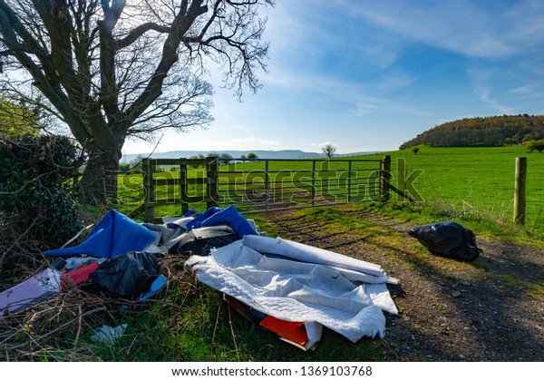 Fly Tipping by A Farmers Field Gate in the\
Countryside with Green Fields Hills in the distance with Trees and\
Light Blue Sky and Shadows