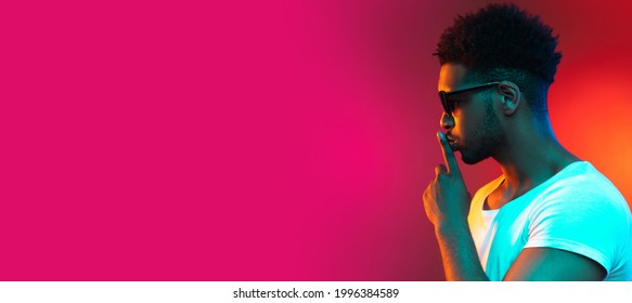 Flyer with portrait in profile of young serious african man in sunglasses standing with his finger to lips on bright pink neon background. Side view.