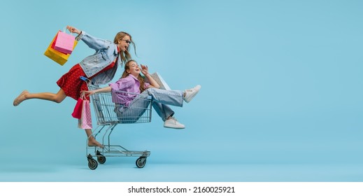 Flyer with pretty happy girls with shopping bags ride on a cart isolated on blue background. Concept of sales, black friday, discount, emotions, youth and fashion. Copy space for ad, text