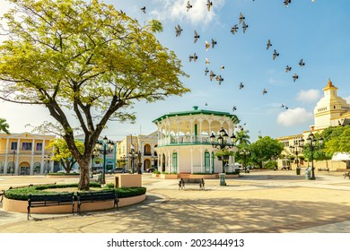 Flying Birds In The Sky Of The Central Park In Puerto Plata, Independence Square, Plaza De Independencia, And A Catholic Church In The Downtown 