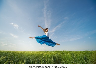 Flying Dancer In The Air. Happy Woman Ballerina In Blue Fabric Skirt Making A Big Jump On Green Field. Summer Or Spring Concept