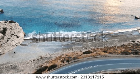 Flying drone over the coastline of the island overlooking the road, rocky shoreline with vegetation and rocks and clear sea with light foamy waves in Cyprus.