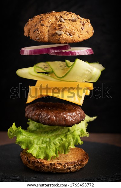 Flying layers of burger with beef\
meet, cheese, cucumber and onion rings against dark\
background