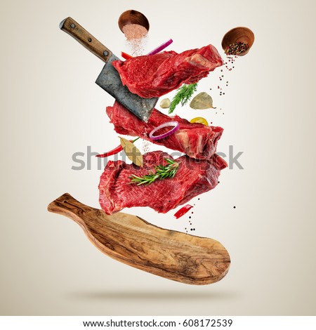 Flying pieces of raw beef steaks, with herbs, served on wooden
board. Meat chopper cutting the flesh. Concept of food preparation in low gravity mode. Separated on smooth gray background