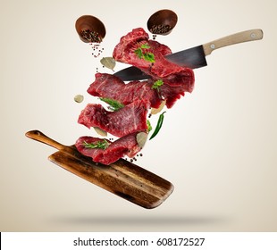 Flying Pieces Of Raw Steaks, With Ingredients For Cooking, Served On Wooden Board. Knife Cutting The Meat. Concept Of Food Preparation In Low Gravity Mode. Separated On Smooth Background