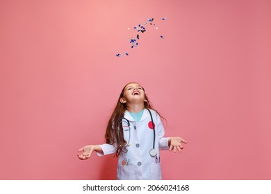 Flying Pills. Studio Shot Of Beautiful Little Girl, Child Playing Doctor Wearing White Lab Coat Posing Isolated On Pink Studio Background. Concept Of Childhood, Studying, Leisure Games.