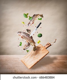 Flying Raw Whole Trout Fishes With Vegetables, Oil And Spices Ingredients Above Wooden Cutting Board For Tasty Cooking On Desk Kitchen Table At Beige Background. Healthy Food Or Diet Eating  Concept.