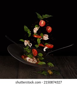 flying salad of mozzarella cheese, basil leaves, ripe cherry tomatoes, green olives, spices with cutlery, on black dishes and black background - Shutterstock ID 2150874327
