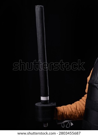 Foam-padded training sword held by fencer wearing protective jacket on black backdrop.