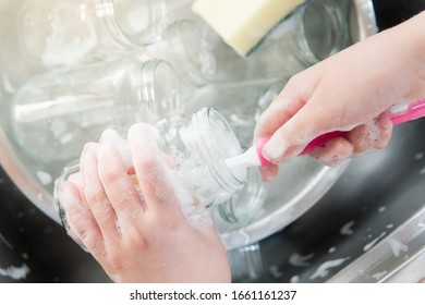 Foamy Hands Of Child Wash & Clean Glass Jars In Kitchen Sink With Bottle Brush. Daily Chores Improve Life Skills, Growth Mindset, Self-confidence, Responsibility. Montessori Child Development Concept.