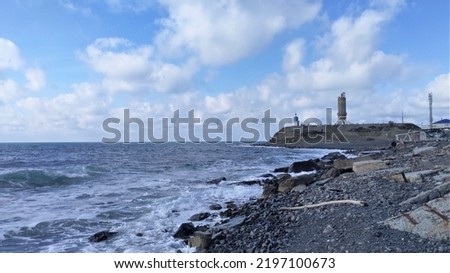 Foamy sea waves roll on the rocky shore. On a hilly promontory stand a lighthouse, a chapel and a metal communication tower. The sky above the sea is blue with clouds