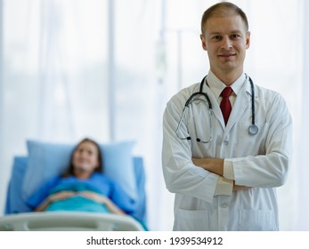 Focus to caucasian doctor man wearing uniform carrying stethoscope on neck smiling and standing folded crossed arms with background are woman patient sleep on bed in room at hospital in the morning.
