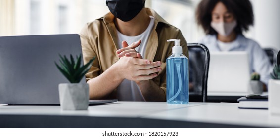 Focus On Antiseptic To Protect Against Covid Virus. Guy Disinfects His Hands, Sitting At Table At Workplace With Laptop, Working In Interior Of Modern Office, Panorama, Copy Space