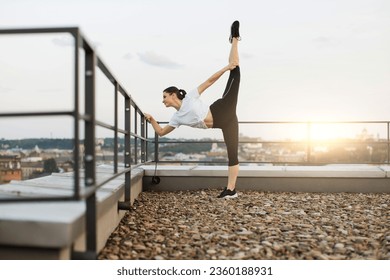 Focus on attractive brunette woman in activewear lifting left leg high to sky while holding handrails on terrace. Slender young person performing standing split during outdoor physical activity. - Powered by Shutterstock