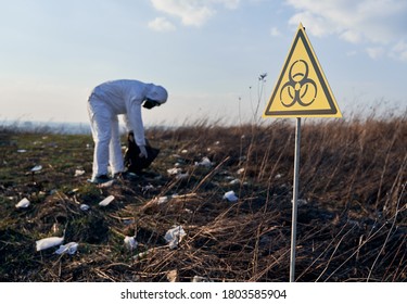 Focus On Biohazard Sign. On The Blurred Background Environmentalist Wearing White Protective Suit, Gas Mask, Collecting Plastic Garbage Into Black Waste Bag Outdoors On A Sunny Day. Concept Of Ecology