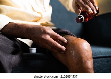 Focus On Bottle, Close Up Of Unrecognizable Old Man Using Ayurvedic Oil Massage For Knee Joint Paint At Home Before Sleeping - Concept Of Using Natural Pain Relief Oil For Osteoarthritis