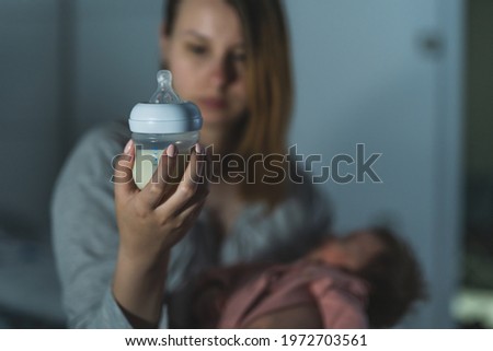 Focus on bottle milk instant formula mother checking content while holding her newborn baby sitting on bed at night in dark room at home