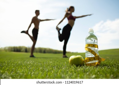 Focus on bottle of water with centimeter band and apple on grass. Sporty couple is doing stretching exercises together on lawn. They are standing on one foot and bending other while keeping balance