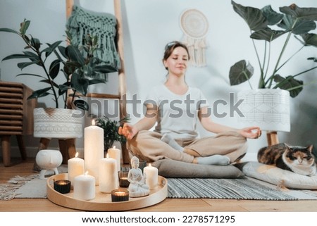 Focus on burning candles and white Buddha statuette on tray with background of woman practicing meditation at home with cat in modern Scandi interior. Zen Composition for yoga practice, relaxation.