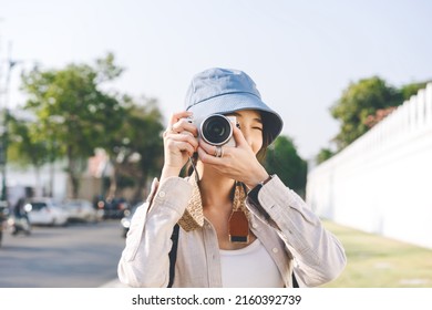 Focus on camera. Young adult asian woman traveller wear blue hat and backpack for photo trip. People traveling in city lifestyle on day. Staycation summer trip concept. - Powered by Shutterstock