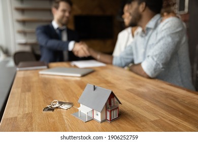 Focus On Close Up Keys Bunch And Cottage House Toy Model On Wooden Table. Smiling Young Man Broker Realtor Real Estate Agent Shake Hands Of Happy Black Couple Clients Homeowners On Blurred Background