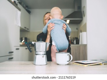 Focus On Coffee Maker And Cup, Background People Blurred. Modern Young Tired Mom And Little Child After Sleepless Night. Exhausted Woman With Baby Is Sitting With Coffee In Kitchen. 