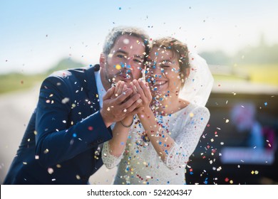 Focus on the confetti. portrait of newlyweds. the groom blowing on confetti and the bride smiling. their hands meet. the focus is on them while their car with just married is blur. shot with flare