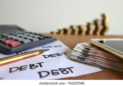 Focus On Debt Word On White Paper And Mobile Phone On Pile Of Money, Calculator, Gold Pen And Decreasing Unstable Rolls Ladder Of Coins On Wood Table In White Background