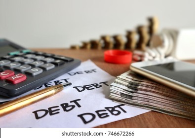 Focus On Debt Word On White Paper And Mobile Phone On Pile Of Money, Calculator, Gold Pen, Decreasing Unstable Rolls Ladder Of Coins And Pills Pouring From Bottle On Wood Table In White Background