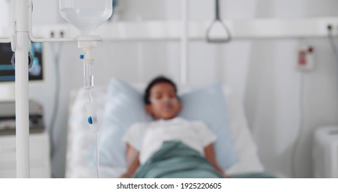 Focus On Drip In Hospital Room With Blurry Patient Child On Hospital Bed. Sick African Little Boy Resting In Bed Having Dropper With Medication In Ward