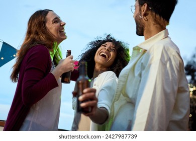 Focus on excited African woman holding beer bottle with laughing group millennial friends at sunset rooftop party. Gathering of cheerful young people enjoying summer day outdoors having fun together - Powered by Shutterstock