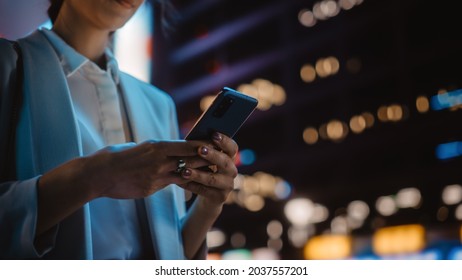 Focus On Female Hand Using Smartphone Walking Through Night City Street Full Of Neon Light. Woman Holding Mobile Phone, Posting Social Media, Online Shopping, Texting.