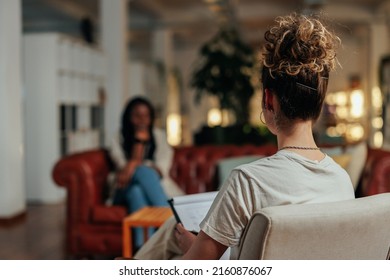 Focus on female psychiatrist writing information of african female patient while sitting on sofa and talking about his illness - Shutterstock ID 2160876067