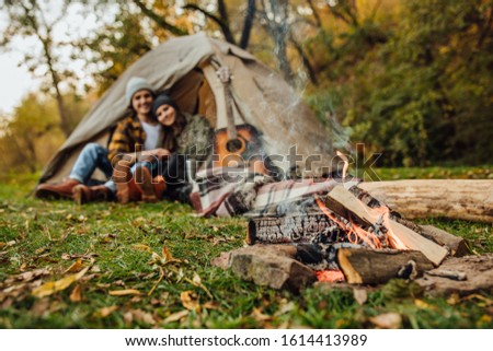 Focus on fire near tent. Young loved couple of tourists have a date in the forest. Attractive young woman and handsome man are spending time together on nature. Hugging lovers near campfire.
