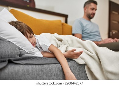 Focus On Foreground Of Little Boy Sleeping Under Blanket Near Blurred Father On Sofa At Home. Young Caucasian Family Relationship. Fatherhood And Parenting. Modern Domestic Lifestyle