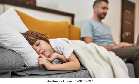 Focus On Foreground Of Little Boy Sleeping Under Blanket Near Blurred Dad Using Laptop On Sofa At Home. Young Caucasian Family Relationship. Fatherhood And Parenting. Modern Domestic Lifestyle