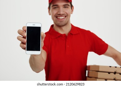 Focus On Foreground Of Smartphone In Hand Of Smiling Caucasian Male Courier Holding Cardboard Boxes. Shipping And Logistics. Partial Image Of Young Man Looking At Camera. White Background In Studio