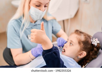 Focus on girl's hand giving thumbs up while woman stomatologist in medical mask sitting beside kid and examining child teeth. Concept of pediatric dentistry and dental care approval.