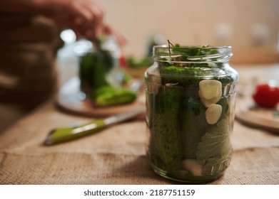 Focus On A Glass Jar, Filled With Freshly Pickled Cucumbers, Herbs And Fresh Garlic Cloves, On A Blurred Background Of A Woman, Housewife Making Pickles At Home Kitchen. Marinating, Canning, Pickling.