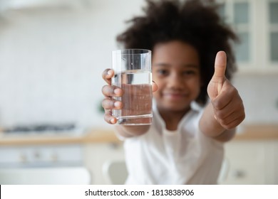 Focus On Happy Small African American Girls Hands Holding Glass With Fresh Pure Water And Showing Thumbs Up Gesture. Smiling Little Biracial Kid Recommending Healthcare Habit, Morning Refreshment.