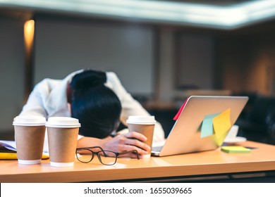 Focus On Hot Coffee Cup And Glasses On Office Desk. Background With Asian Business Woman Exhausted Tried And Sleep Form Work Overtime On Late Night.