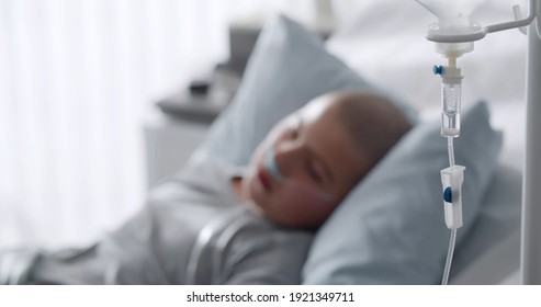 Focus On Intravenous Drip With Sick Kid Lying In Hospital Bed On Background. Teen Boy Patient With Cancer Resting In Bed Having Medication In Dropper