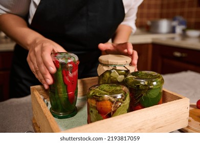 Focus on a jar with canned fermented red and green hot chili, in the hands of a housewife, putting it upside down on a wooden crate with homemade preserved and marinated vegetables. Canning food - Shutterstock ID 2193817059