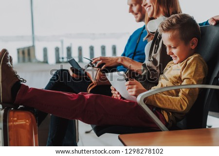 Focus on joyful boy sitting near his parents in lounge of airport. He is putting his feet on suitcase and using touchpad while looking at it