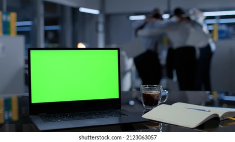 Focus On Laptop With Green Screen On Desk In Office. Business Team Celebrating Success On Blurred Background Late In Office