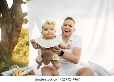 Focus On Little Baby Girl. Beautiful Young Father Lifting Up His Cute Little Daughter On Yellow Dress On Hands While Playing On Boho Picnic. Family Time Outdoors.